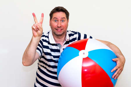 Front view portrait of a surprised middle age guy holding beach ball and showing victory gesture on white studio wall.の写真素材