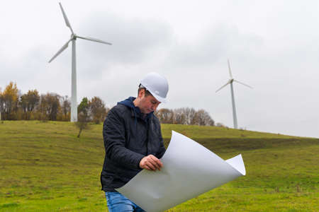 Side view portrait of one male engineer work by blueprint at the wind energy field autumn cloudy sky...
