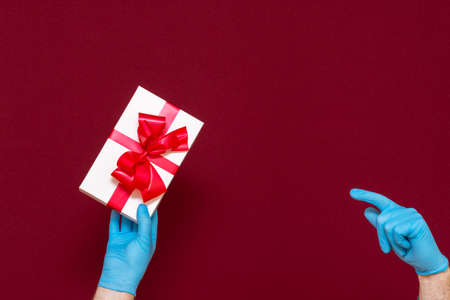 New Year's 2021. Christmas celebration, protection from coronavirus. Man hands wearing medical rubber blue gloves pointing the present gift box.Red studio background.の写真素材