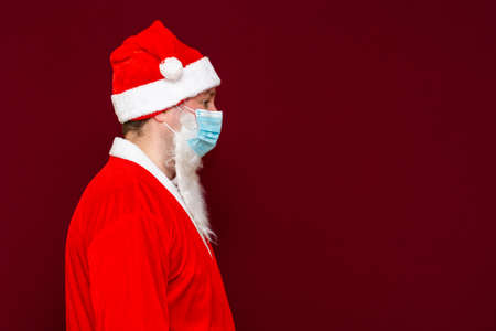 Side view Santa Claus man wears face mask safe from coronavirus virus covid-19 during quarantine on red studio background.Close up.の写真素材