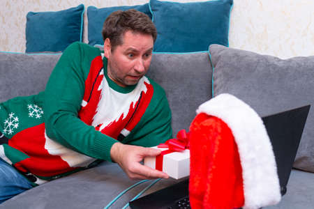 Male man virtually exchanging Christmas gifts and having video call over a laptop at home on the couch.の写真素材