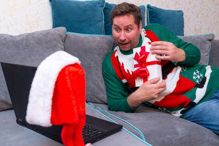 Male man virtually exchanging Christmas gifts and having video call over a laptop at home on the couch.の写真素材