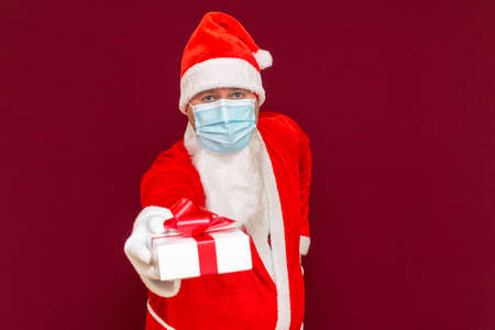 Christmas gift. Santa Claus wears medical mask holding gift box and looking at camera while standing against red studio backgroundの写真素材