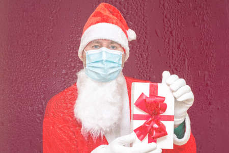 Christmas gift. Santa Claus wears medical mask holding gift box and looking at camera while standing...