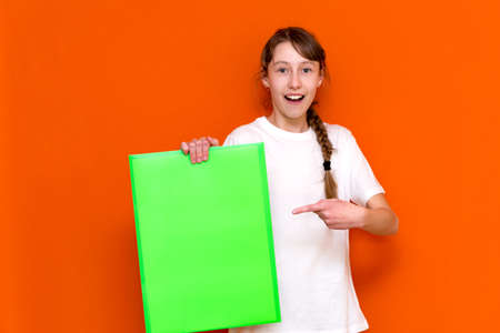 Surprised caucasian young girl poke at folder with a banner advertisement on a orange studio background. copy space bannerの写真素材