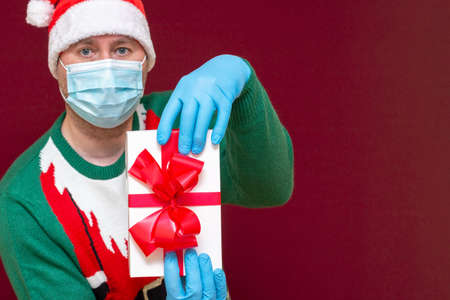 Closeup Handsome male wears Santa hat medical mask gloves holds gift box in hands and showing on camera red studio background.Selective focus.の写真素材