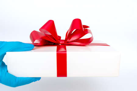 Hand wearing meidical gloves holding gift box with red ribbon. Pandemic protection.Bright white background.の写真素材
