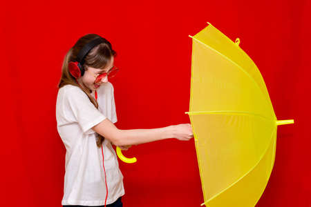 Teenage girl listens to music in headphones with umbrella. red studio background wearing white t...