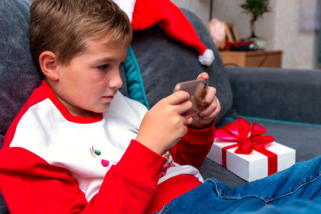 Closeup Attractive kid boy using smartphone on Christmas holidays white gift for xmas photo at home. Side view image.の写真素材