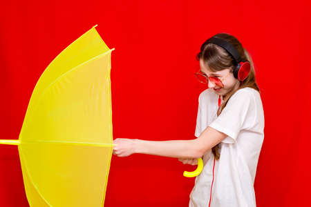 Teenage girl listens to music in headphones with umbrella. red studio background wearing white t shirt.studio shot.の写真素材