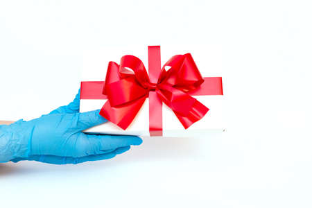 Holiday Christmas gift. COVID-19 prevention.Hands wearing meidical gloves holding gift box with red ribbon. Pandemic protection.Bright white background.の写真素材