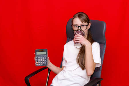 Young surprised caucasian girl counting calculator drinks coffee. Education concept.Red studio background.の写真素材