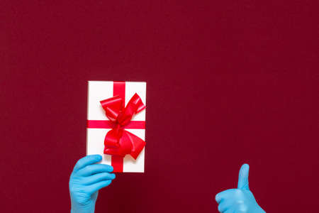 Man hands wearing medical rubber gloves holding present gift box,showing thumb up.Red studio background.の写真素材