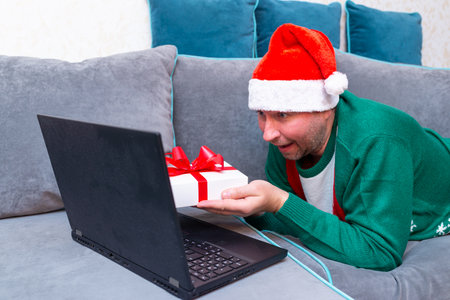 Male man virtually exchanging Christmas gifts and having video call over a laptop at home on the couch.の写真素材