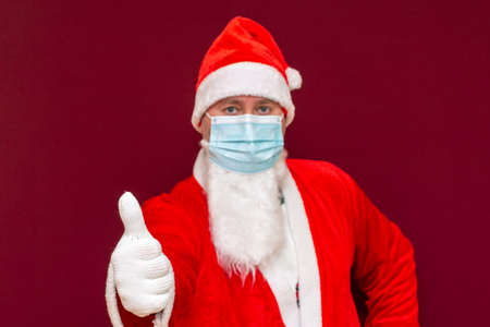 Closeup Santa Claus wearing medical mask shows thumb up New year and Merry Christmas and happy holidays concept.Red background.Selective focus.の写真素材