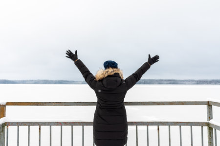 Woman spread her arms out in warm clothes down jacket standing in the snow back and admires the beautiful landscape with frozen lake with a thick layer of snow. Foggy day.Back view.の写真素材