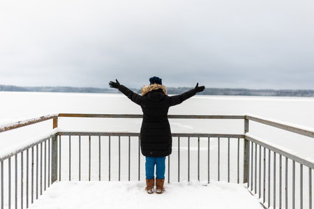 Woman spread her arms out in warm clothes down jacket standing in the snow back and admires the beautiful landscape with frozen lake with a thick layer of snow. Foggy day.Back view.の写真素材