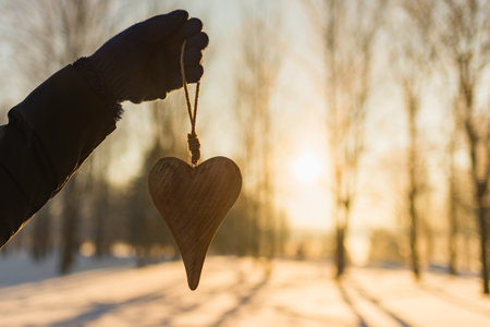 Womans hand holding a wooden heart on winter snowy forest evening blurred background. Love winter or St. Valentine's day concept. Selective focus.の写真素材