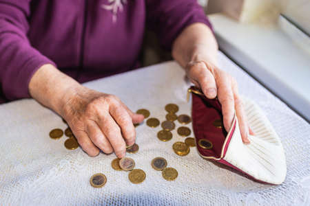 Concerned elderly woman sitting at home and counting remaining coins.old woman sitting miserably at the table at home and counting remaining coins from the pension in her wallet after paying the billsの写真素材