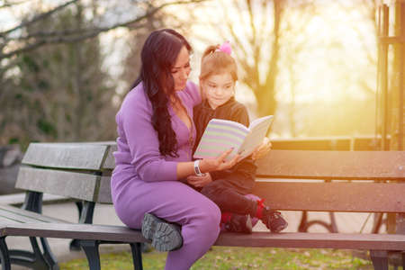 Beautiful little girl reading book with her mother and smiling. Summer blurred park in background.woman reading a book to her daughter enjoying a warm evening in the local sunset parkの写真素材