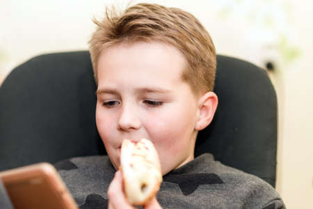A Hungry boy eating a hot dog at home sit while playing with the smartphone. Indoors shot.Closeup.