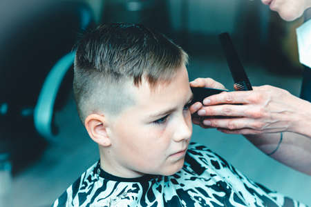 Hairdresser makes a fashionable pretty hairstyle for the young boy in a modern barbershop.Side view.Toned.Closeup.の写真素材