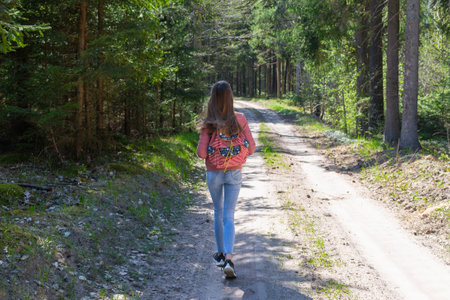 One young girl tourist with backpack walking along path through forest during summer day. Hipster...