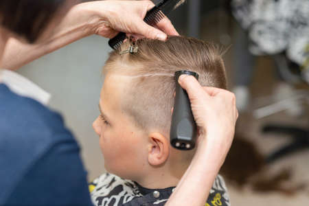 Hairdresser makes a fashionable pretty hairstyle for the young boy in a modern barbershop.Side view.Closeup.の写真素材