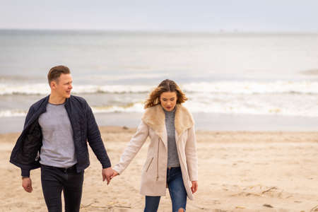 Attractive young couple walking along the shore of a sandy beach, on a spring romantic holiday, outdoors. Travel tourists lifestyle. Couple enjoying loving time together.の写真素材