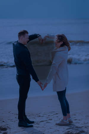 young couple making heart shape with arms on sea beach against dark moonlight tone spring,autumn...
