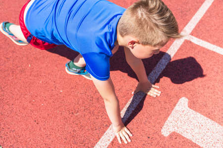 Caucasian schoolboy prepare to start running on a red track.young boy in starting position ready for running. Healthy lifestyle.Summer day.の写真素材