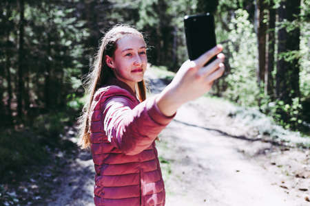 Young teen girl traveler taking selfie on mobile phone in a summer day forest. Smiling girl looking at smartphone camera.Nice spring sun shine day.Selective focus.の写真素材