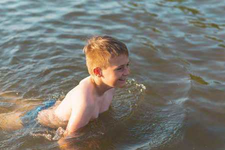 Cute young boy enjoy having fun at lake or beach water on warm sunset evening time outdoors.Happy childhood vacation at countryside concepton.Boy at pier warm summer day having good time in a water.の写真素材