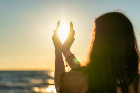 Nice summer sun solstice concept. Silhouette of young woman's hands relaxing, happy meditating and holding sunset against golden hour sky on the beach with natural ocean, sea background.の写真素材