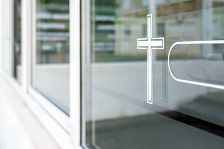 Closeup of a white cross on a window. church in the background.Funeral house summer day after...