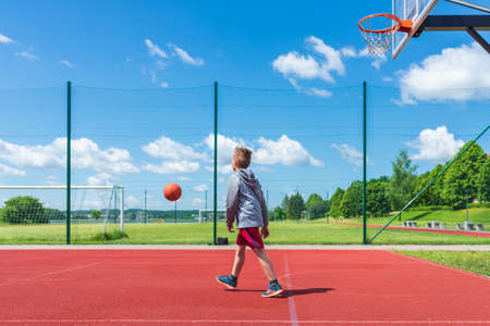 Joyful young boy kid playing basketball on playground.Boy performs shot at red court ball game on the playground during summer day.Sunny urban day.の写真素材