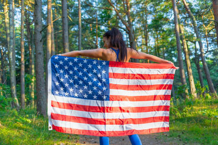 Beautiful girl with American flag.Sexy caucasian girl with American flag in hands on warm summer forest.Back view.の写真素材