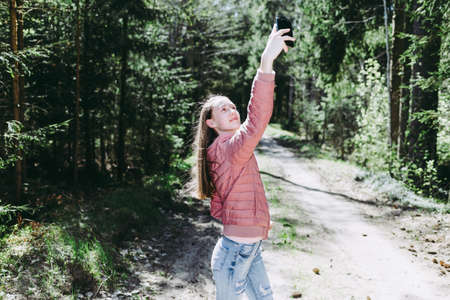 Young teen girl traveler taking selfie on mobile phone in a summer day forest. Smiling girl looking at smartphone camera.Nice spring sun shine day.の写真素材