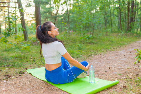 Young attractive slim woman doing yoga in the park. Healthy lifestyle concept.Woman meditating relaxing yoga pose in earphones and listening to music on a summer park or forest.の写真素材