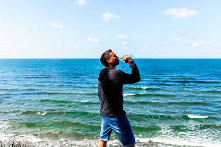 Middle age man drinking water on a sea beach light blue sky clouds. outdoors summer background. Amazing summer travel holiday freedom.Side view.の写真素材