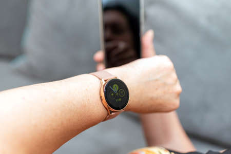 Middle age woman wearing a smartwatch using a mobile phone sitting on a couch in the living room at...