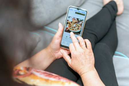 Woman holds mobile phone with a pizzeria website on the screen while lying on a fluffy gray couch....