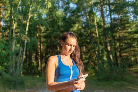 Young pretty female exercising in forest listening to music with earphones. Happy young woman listening to music with headphones in summer forest.の写真素材