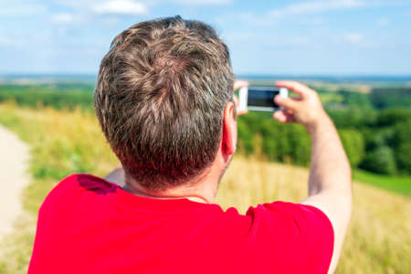 Young male tourist photographs with telephone camera the landscape of the mountain on a camping tour. The man wandering with the help of a phone to make a photo.の写真素材