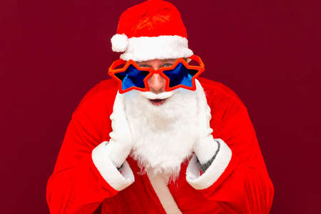 Excited Santa Claus white beard in costume headwear star glasses open mouth staring eyes hear incredible news isolated on red studio background.Closeup.の写真素材