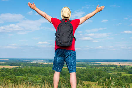Tourist man with backpack enjoying the view in nature.Traveler man with backpack,arms outstretched stand on blue sky white cloud summer landscape day.Man wears hat,red t-shirt blue shorts.Back viewの写真素材