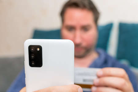 Young man buying fashion items through the Internet on cellphone by using credit card.Online Payment. Frontal portrait of man holding debit card and using smartphone for shopping at homeの写真素材