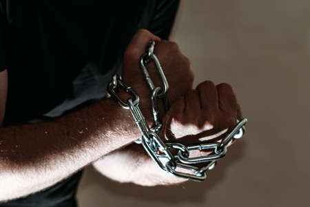 Repented man prisoner with his hands shackled in chains on a dark gray background.Closeup.の写真素材