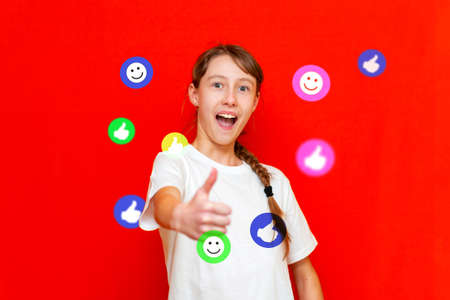 Smiling kid girl 12 years old wearing white t-shirt. Flying thumb up social media services apps around the girl.Showing thumbs up.Red studio background.の写真素材