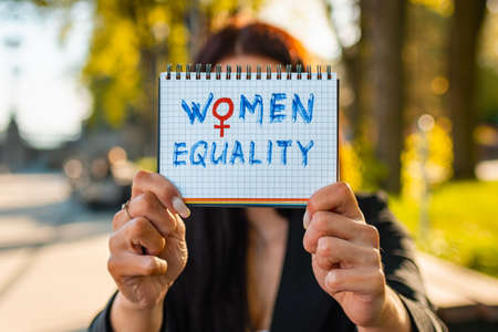 Young caucasian woman outdoors showing a notebook paper in front of her face with the text womens equality written in it.Summer sunset park evening.Closeup.Selective focus.の写真素材